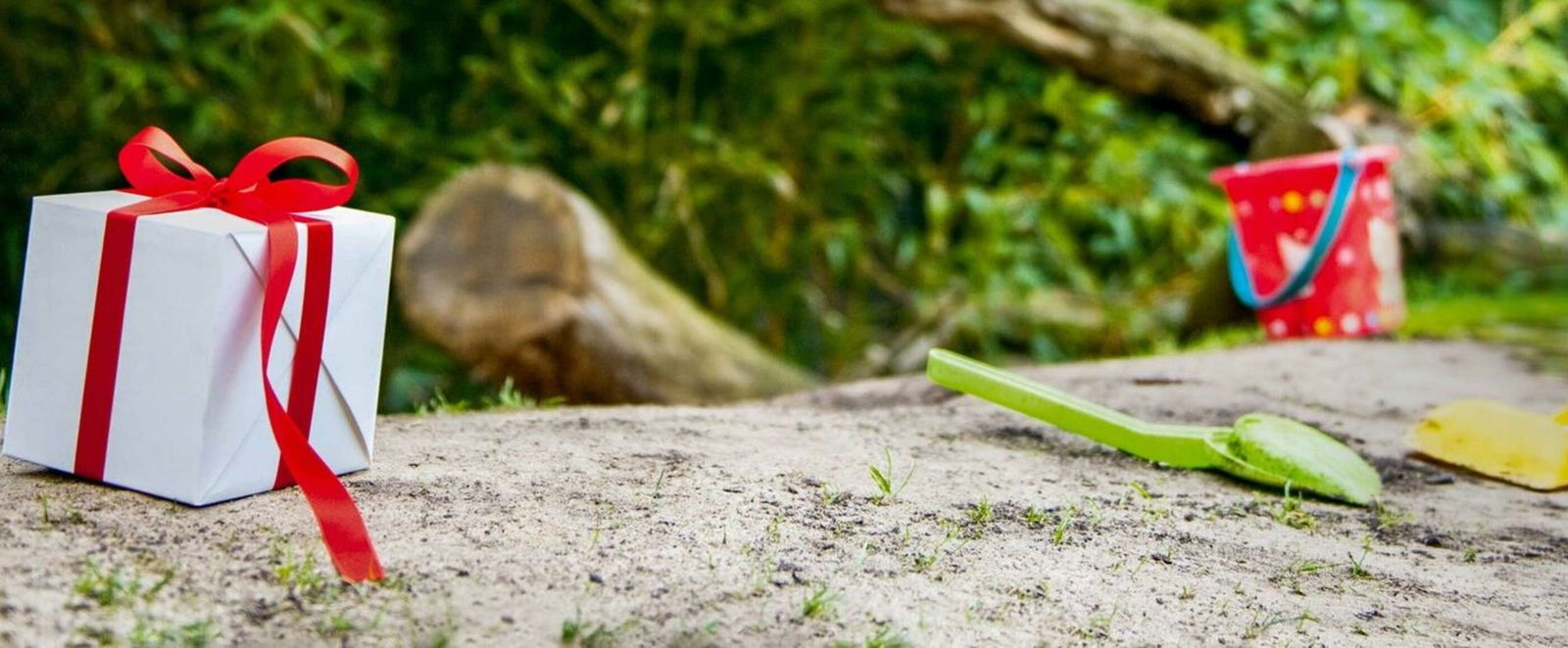 Ein kleines weißes Päckchen mit roter Schleife steht auf Sand, etwas weiter hinten liegen Buddel-Schaufeln für Kinder.