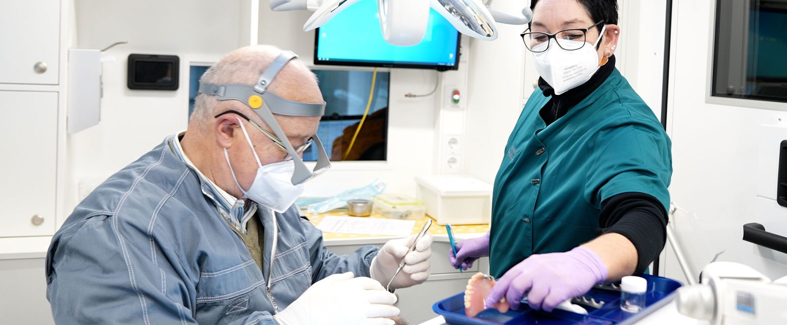 Ein Arzt und eine Arzthelferin, beiden mit Mundschutz und Handschuhen, stehen links und rechts eines Patienten, der in einem Zahnarztstuhl liegt, aber kaum zu sehen ist.