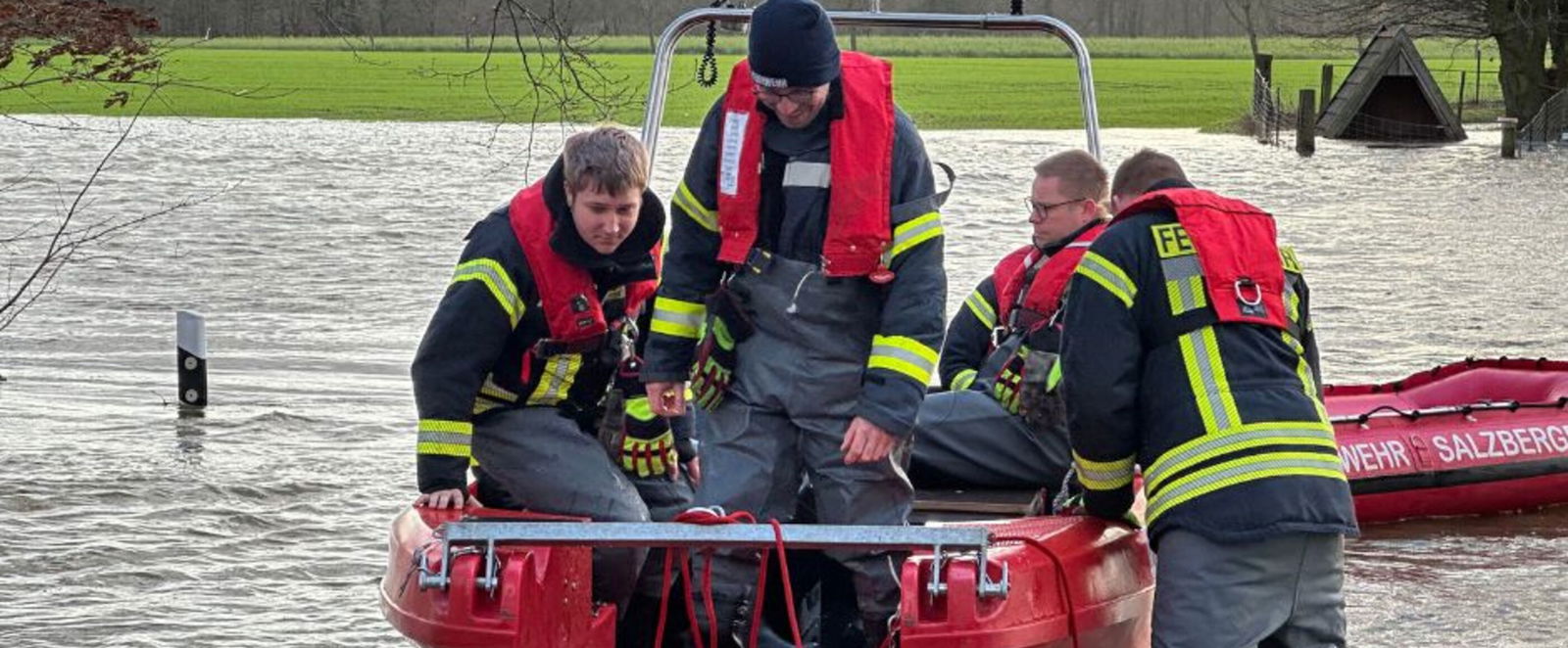 Mehrere Personen mit Feuerwehrjacken sitzen in einem Boot, einer steht draußen im Wasser. Es hat eine Straße überflutet - ein Leitpfosten ragt nur noch ein Stück aus dem Wasser.