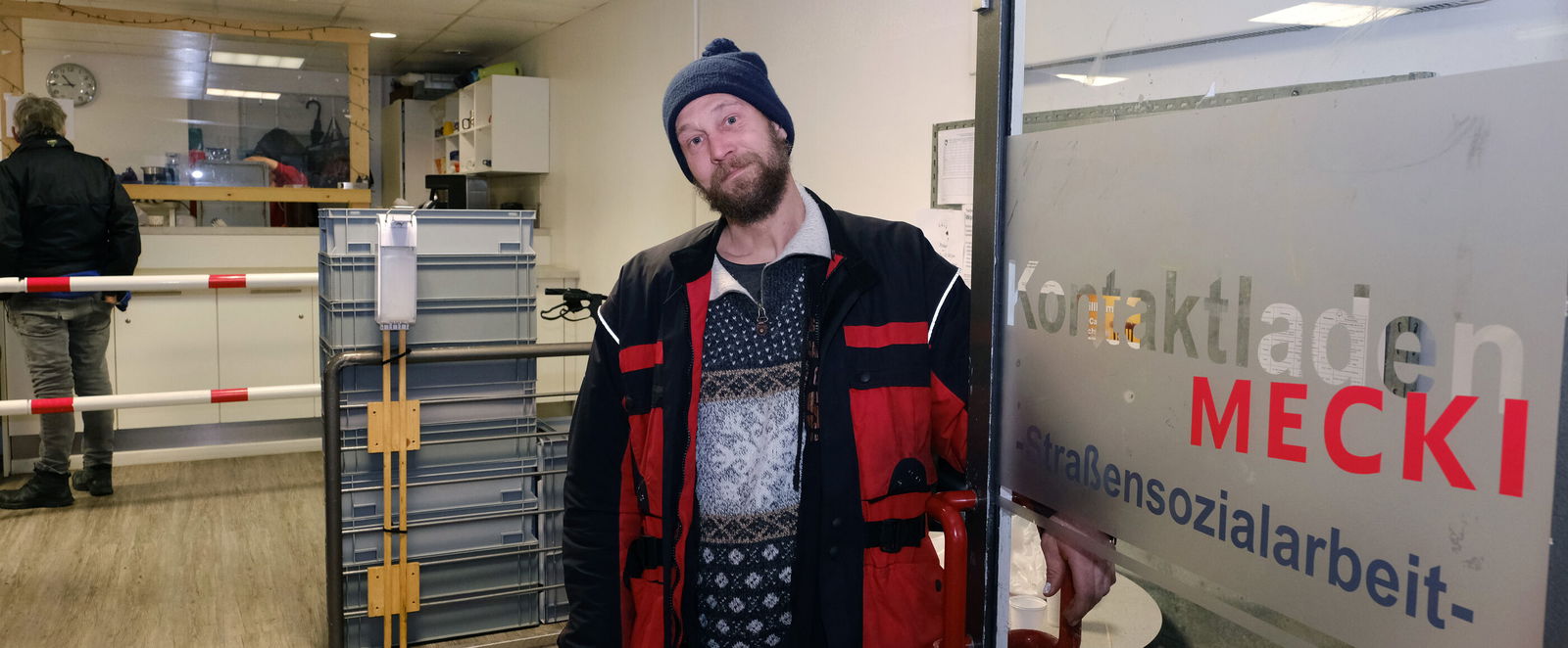 In der geöffneten Glastüre steht ein Mann mit roter Jacke und blauem Winterpulli. Er trägt eine dunkle Mütze und hat braune kurze Haare und einen Vollbart. Er lächelt. Auf der geöffneten Türe steht "Kontaktladen Mecki -Sozialarbeit". Im Hintergrund sieht man eine Essensausgabe. 