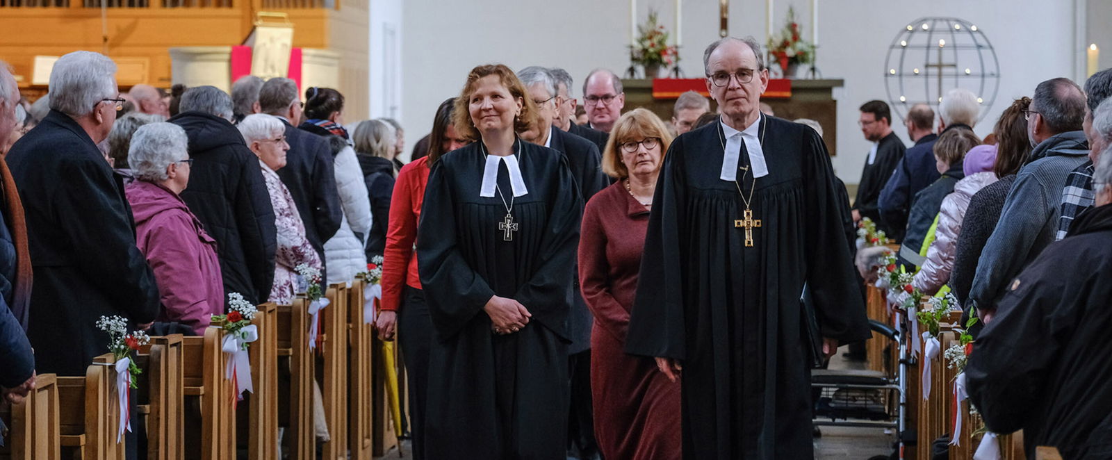Zwei Personen in Talaren gehen zwischen mit Menschen gefüllten Bankreihen in einer Kirche hindurch. Im Hintergrund steht ein Kreuz an einer weißen Wand.