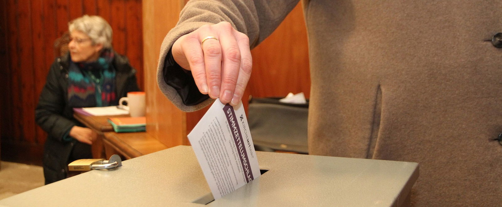 Eine Hand wirft einen Zettel in eine Urne. 