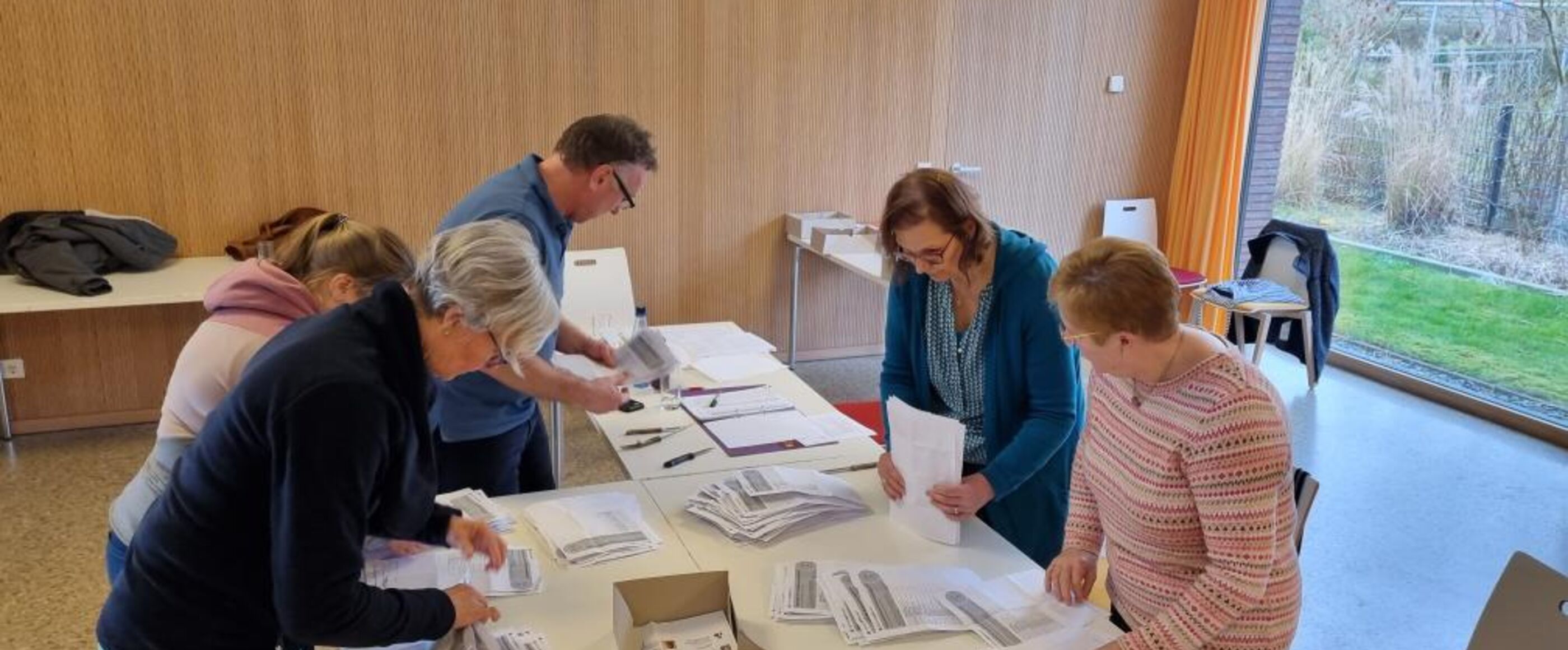 Fünf weiblich gelesene Personen beugen sich über einen Tisch, auf dem Zettel in verschiedenen Stapeln liegen.