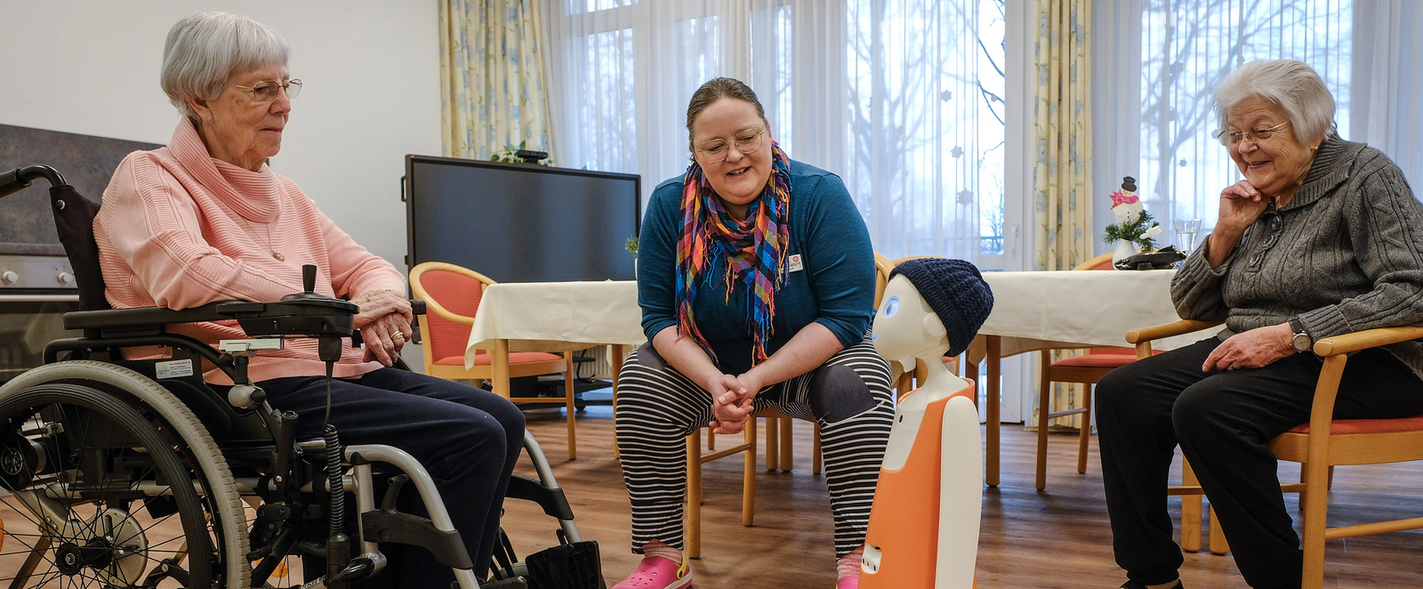 Drei weiblich gelesene Personen sitzen im Halbkreis, eine von ihnen im Rollstuhl. In ihrer Mitte steht ein Roboter, der einem Jungen nachempfunden ist, mit orangenen Hosenanzug und blauer Mütze. Statt Beinen hat er Räder.