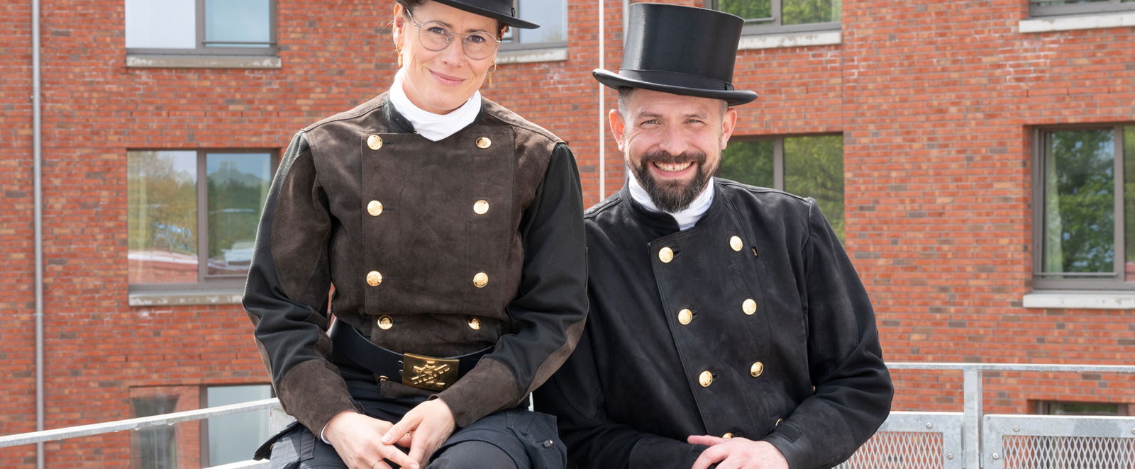 Eine als Frau lesbare Schornsteinfegerin und ein als Mann lesbarer Schornsteinfeger stehen auf einem Dach.