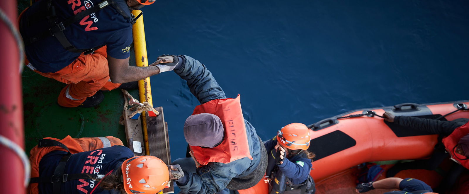 Blick von oben: Zwei Menschen auf einem Schiff helfen einem anderen aus einem Schlauchboot hinauf.