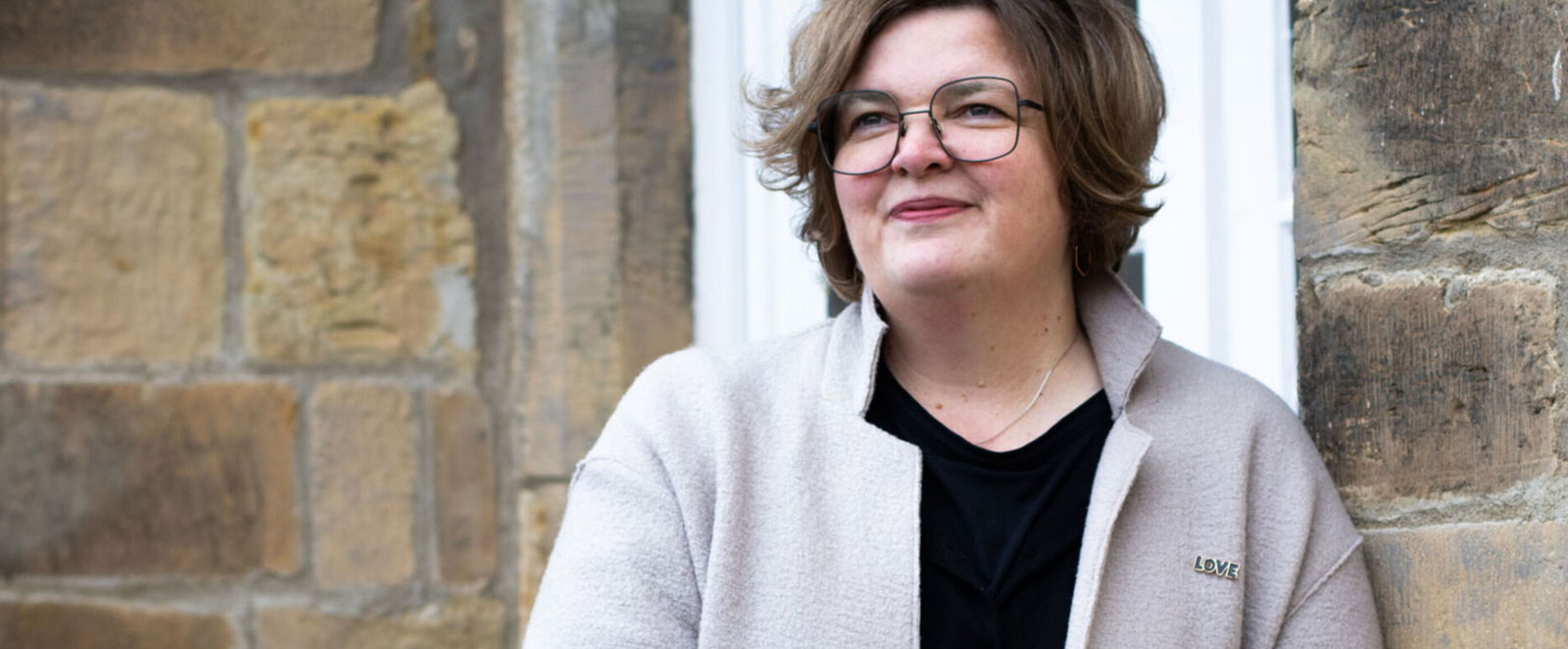 Eine als Frau lesbare Person lehnt an einer Hauswand.