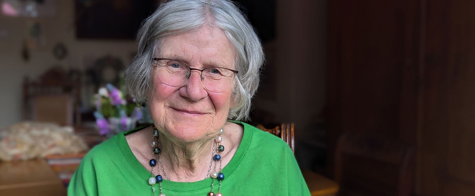 Eine ältere, als Frau lesbare Person im grünen Blümchenpullover sitzt in einem Wohnzimmer.