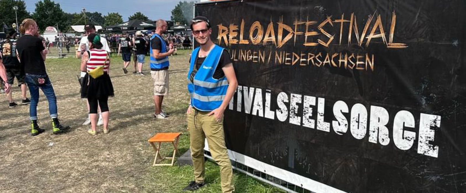 Ein junger Mann steht in einer blauen Wester vor einem Banner auf dem Festivalseelsorge steht.