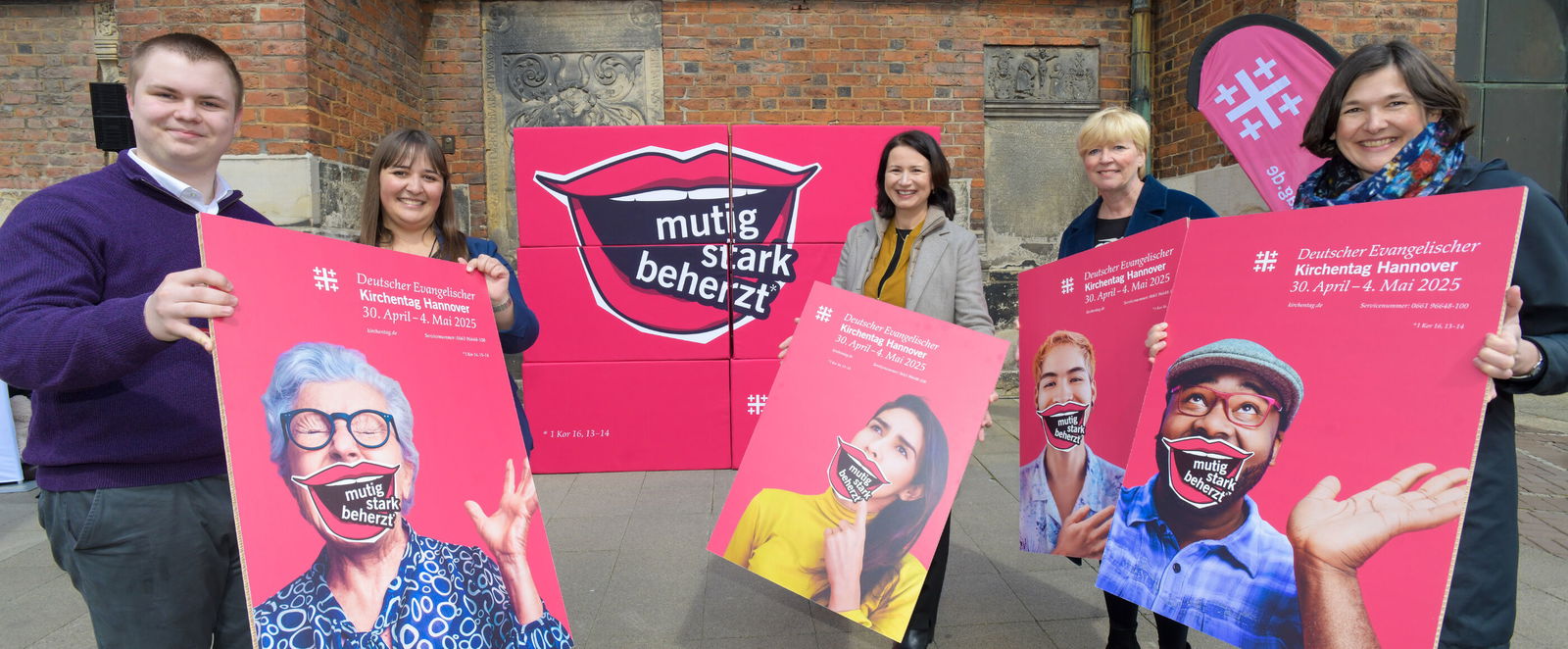 Mehrere als Männer und Frauen lesbare Personen halten Plakate mit Kirchentagswerbung.