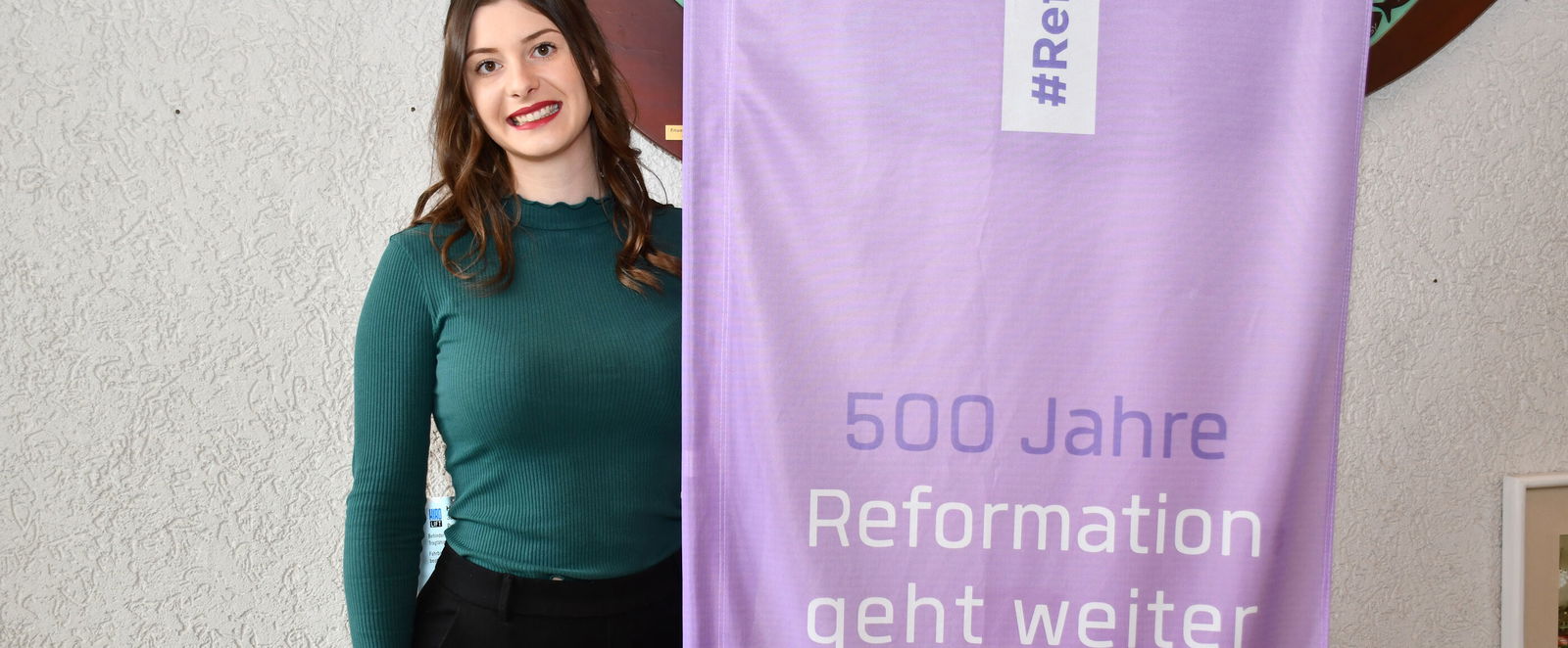Eine als junge Frau lesbare Person steht neben einem lila Banner mit der Aufschrift "500 Jahre - Reformation geht weiter"