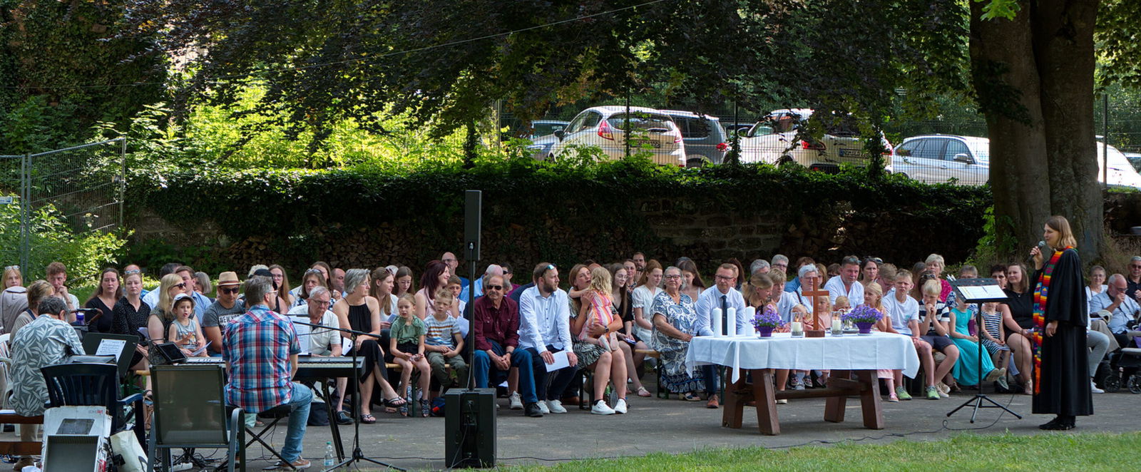 Viele Menschen sitzen im Freien, rechts steht eine Person im Talar, weiter links ein Altar.