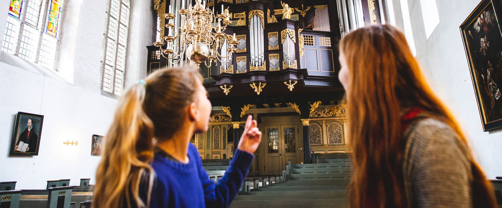 Zwei weiblich gelesene Personen stehen in einer Kirche und deuten auf eine Orgel