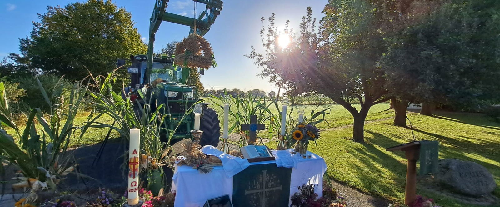 Ein Trecker steht hinter einem Tisch mit Gaben, einer weißen Tischdecke, einer Kerze als Altar. Dahinter sind ein Feld und Bäume erkennbar, die Sonne taucht alles in herbstliches Licht.