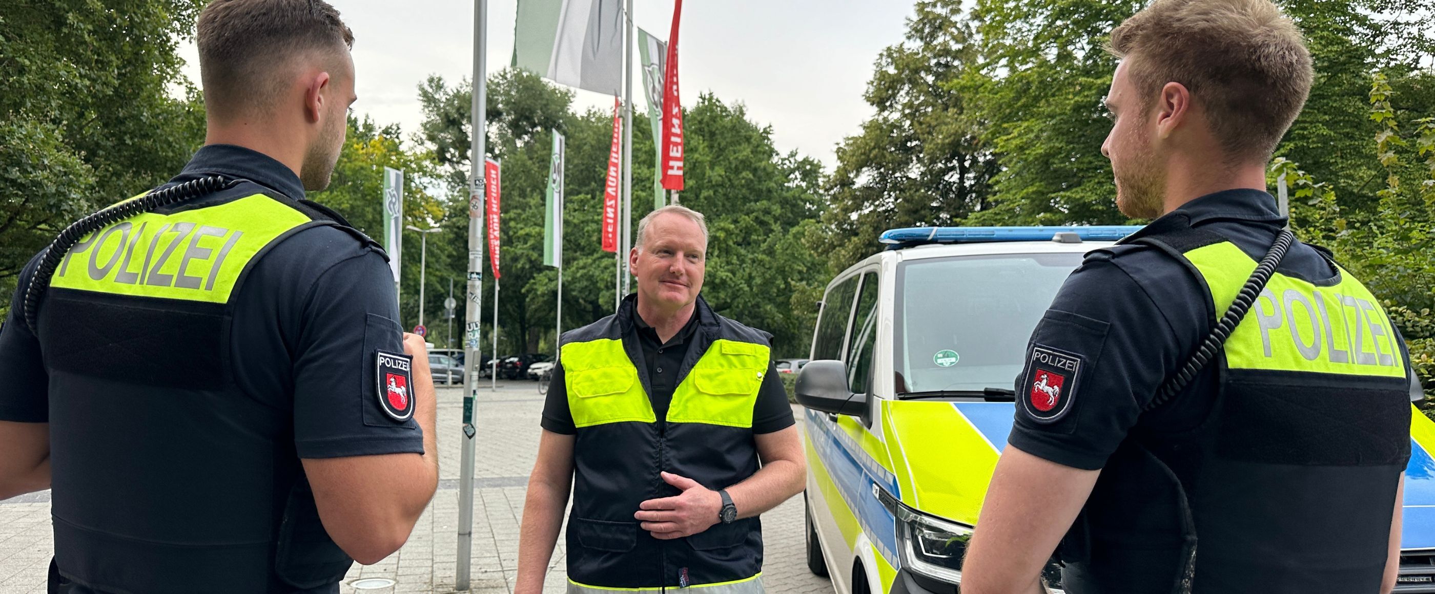 Drei männlich gelesene Personen stehen in Polizeiuniform vor einem Streifenwagen