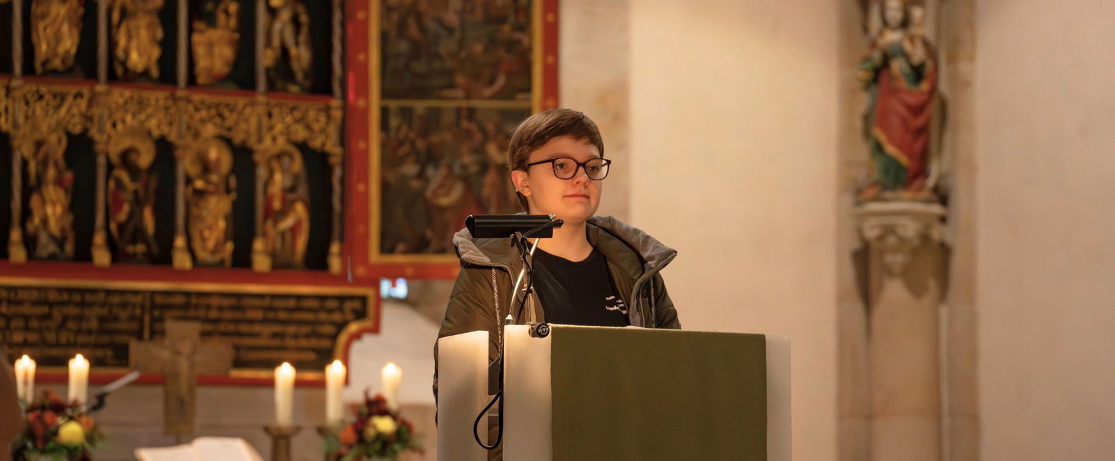 Ein Mädchen mit kurzen Haaren und einer Brille steht vor einem Altar an einem Rednerpult.