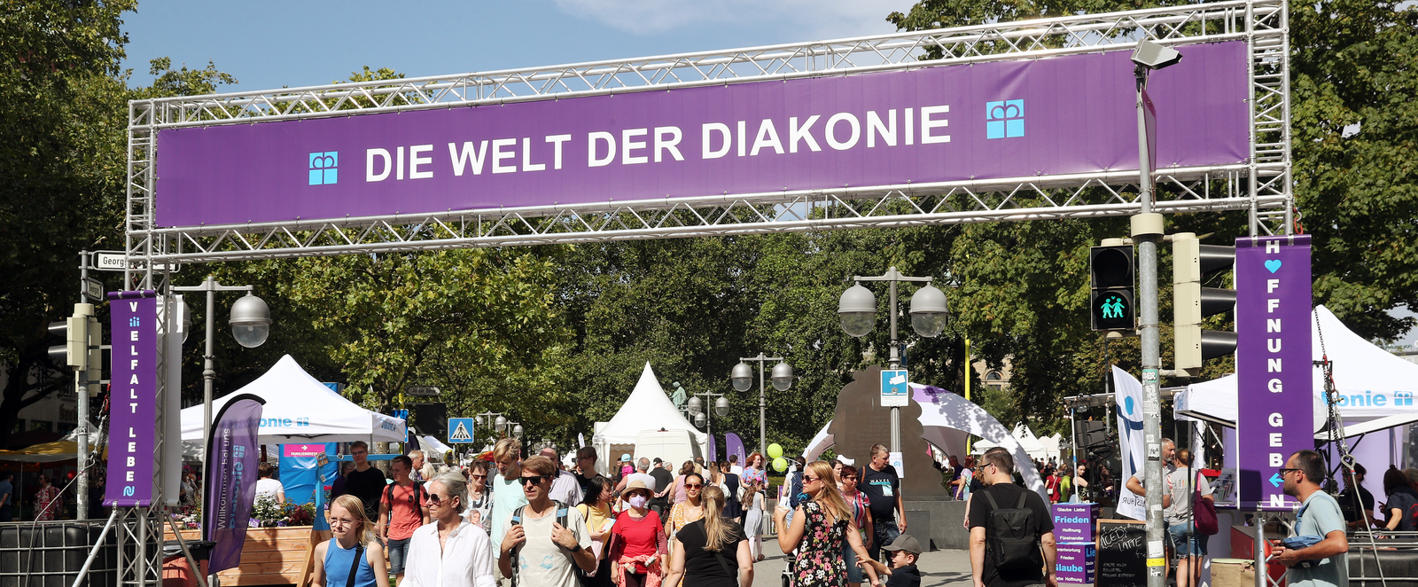 Auf einem großen lila Banner steht: Welt der Diakonie. Menschen in sommerlicher Kleidung gehen darunter hindurch.