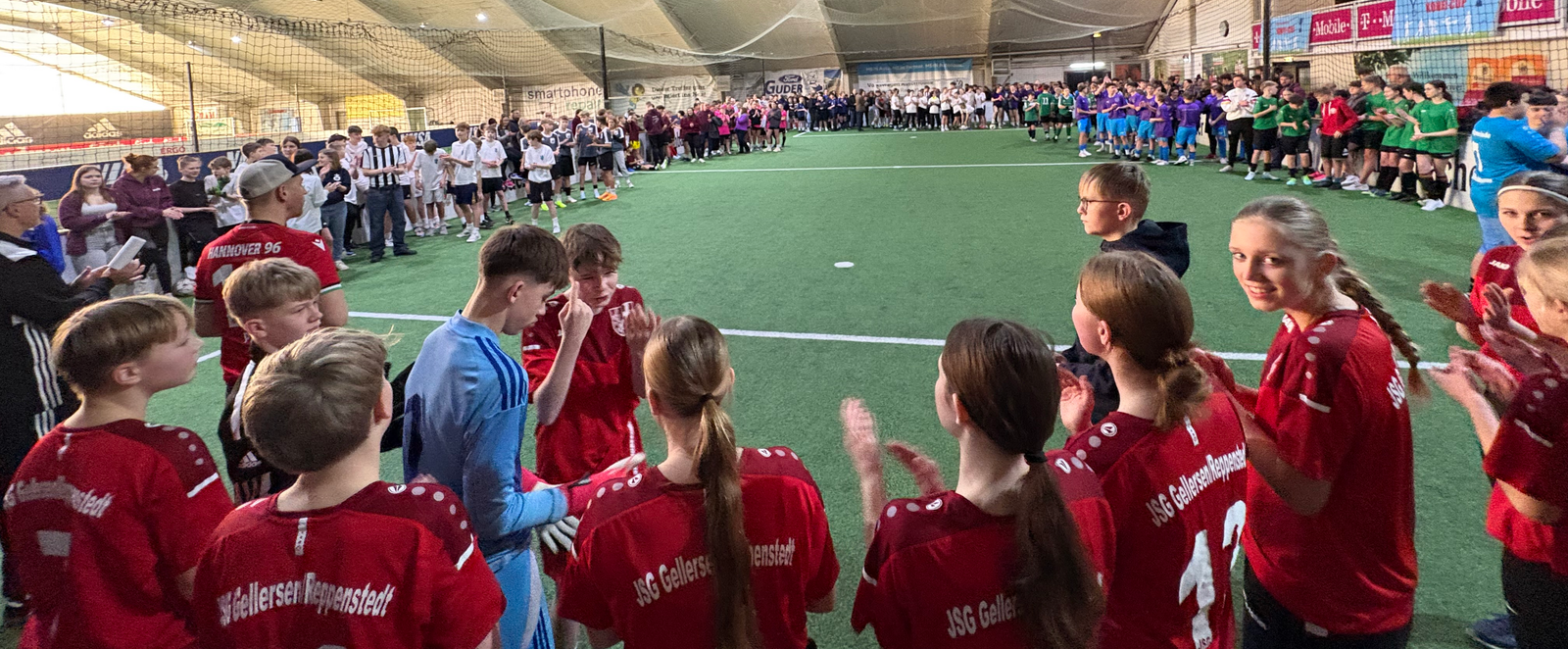 Blick in eine Halle mit Fußballfeldern, junge Menschen in unterschiedlichen Trikots stehen um ein Feld herum.