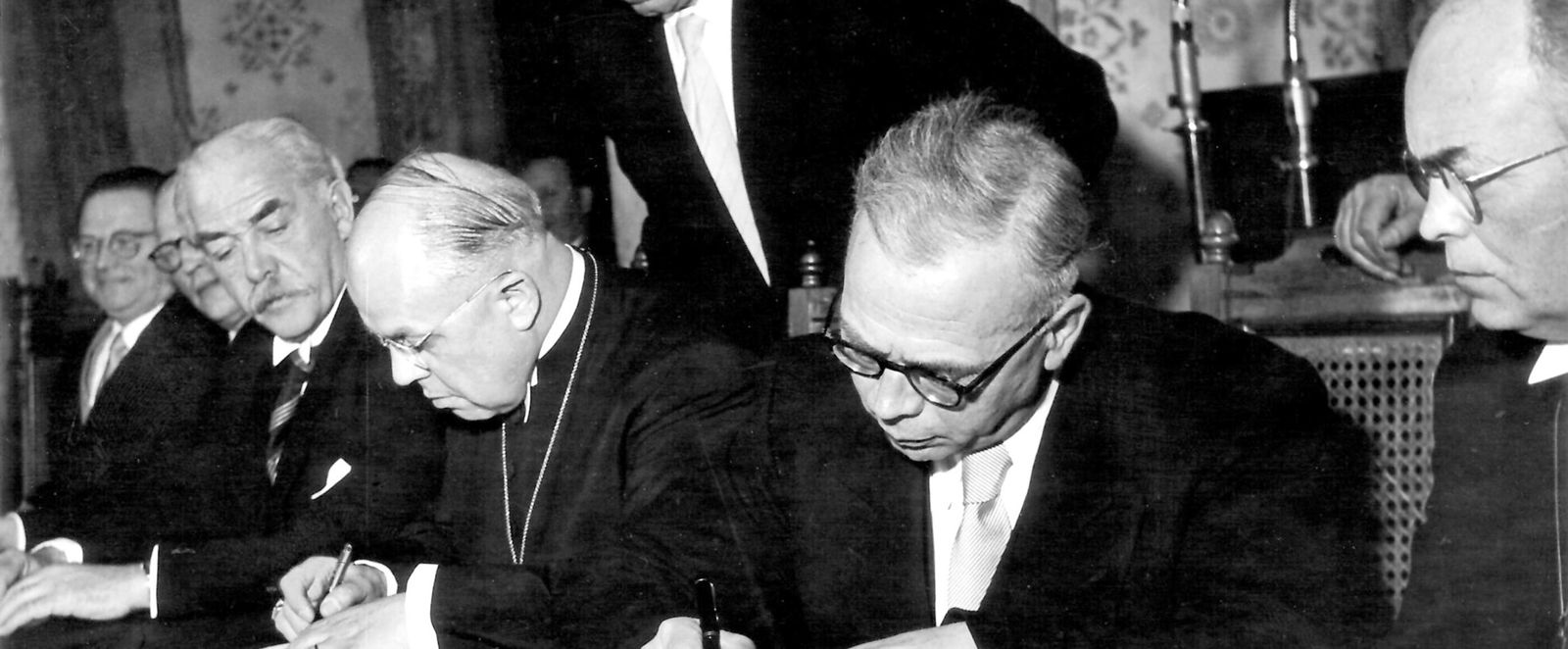 In schwarz-weiß: Sechs männlich gelesene Personen in Anzüge sitzen in Reihe an einem Tisch und unterschreiben Dokumente. Hinter ihnen steht eine weitere Person.