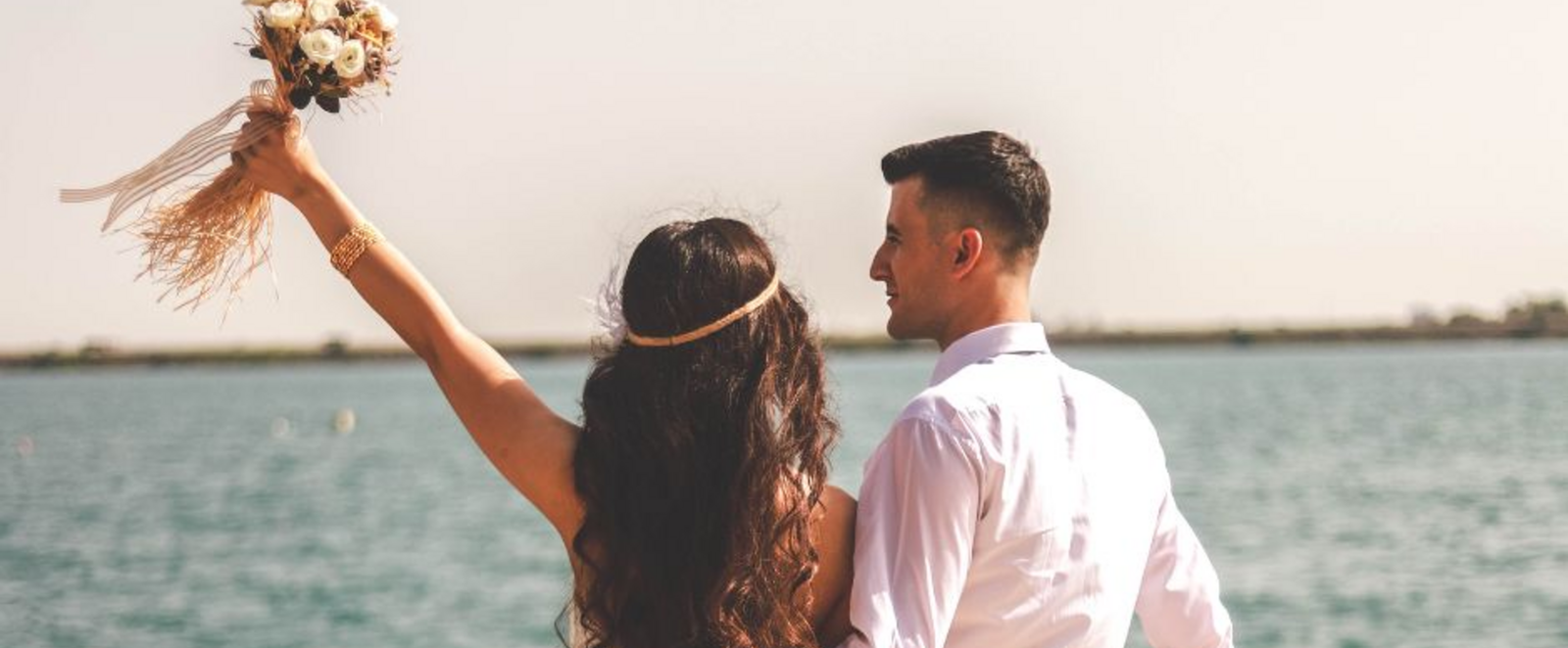 Eine weiblich und eine männlich gelesene Person in Hochzeitsoutfits auf einem Boot auf dem Wasser von hinten zu sehen. Die eine Person reckt einen Blumenstrauß in die Höhe.