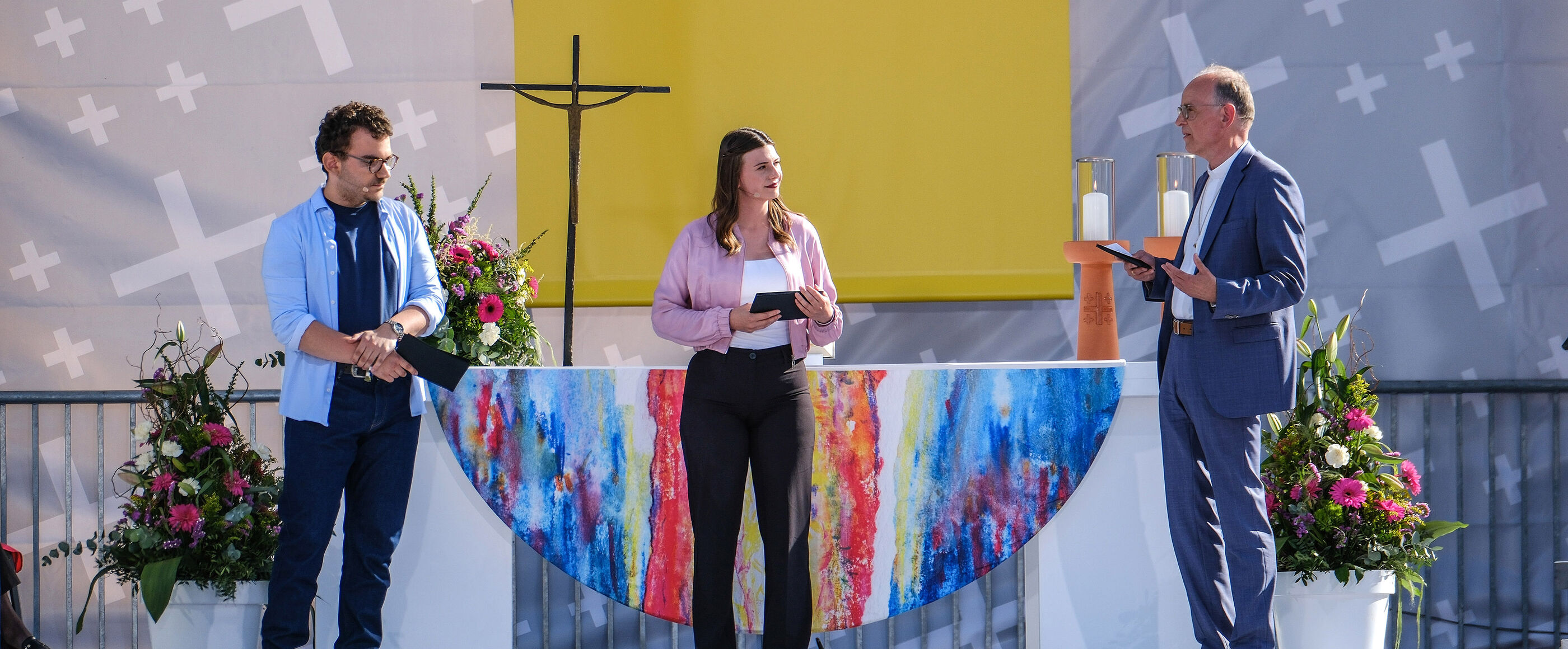 Zwei männlich und eine weiblich zu lesende Person stehen vor einem Altar und unterhalten sich während sie in Mikrofone sprechen.