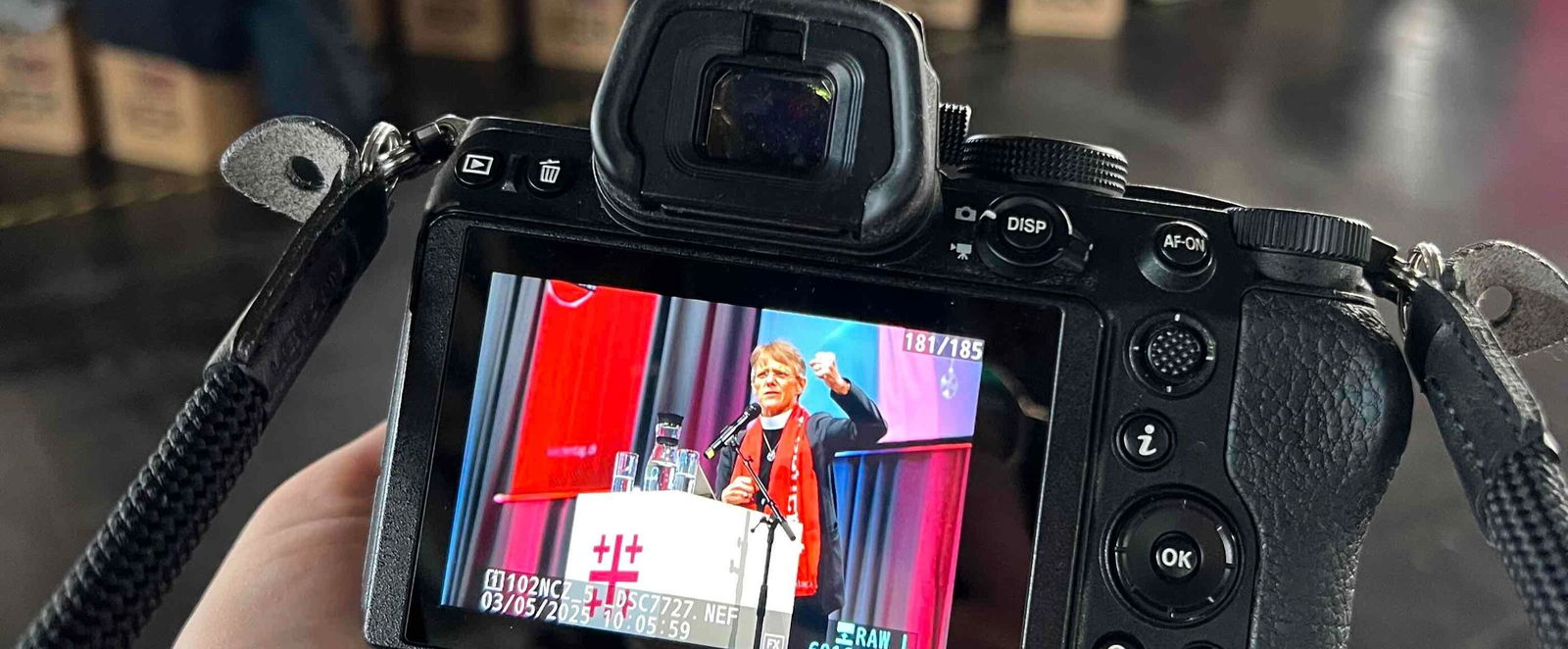 Auf der Anzeige einer Kamera ist eine weiblich gelesene Frau zu sehen, die vor vielen Menschen spricht.