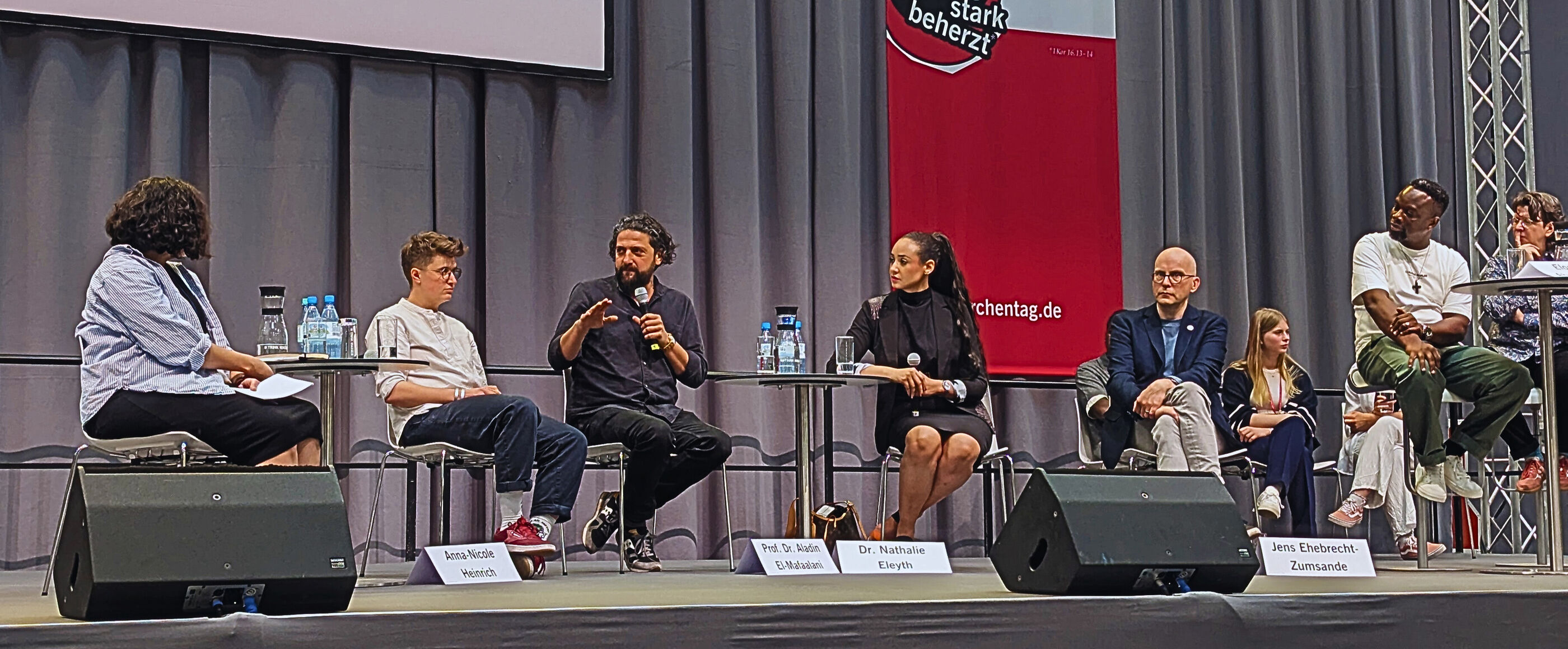 Eine Podiumsdiskussion auf einer Bühne. Hinten hängt eine rote Kirchentags-Fahne.