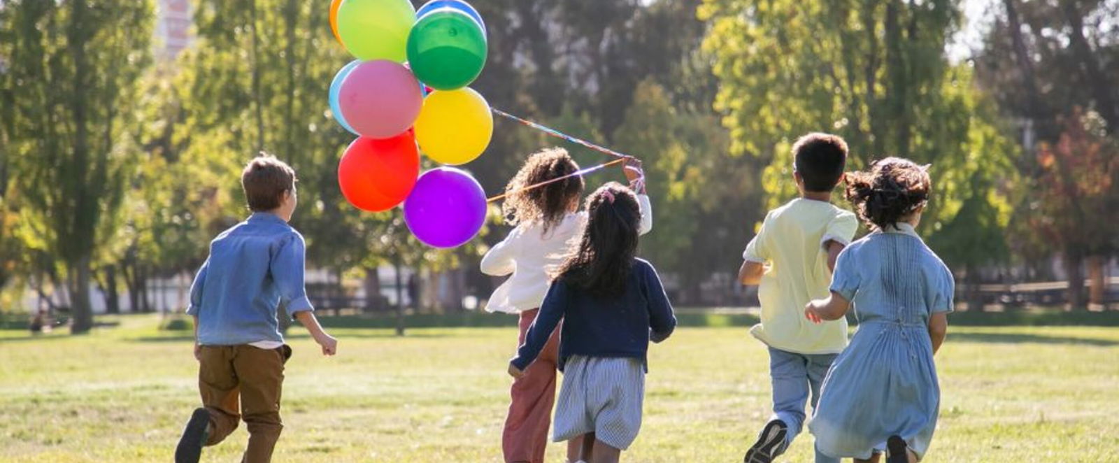Fünf Kinder von hinten, laufen über eine Wiese. Eins zieht Ballons an Schnüren hinter sich durch die Luft.