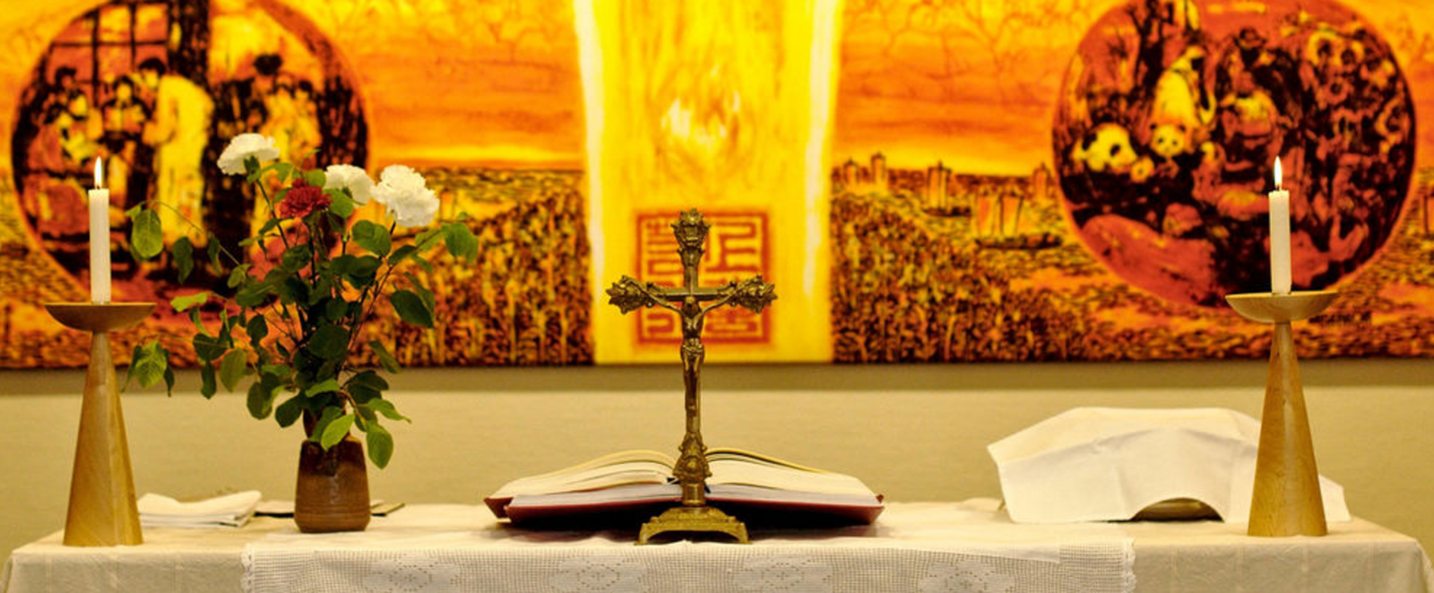 Vor einem leuchtend gelb-orangenen Hintergrund stehen ein Altarkreuz und Blumen.