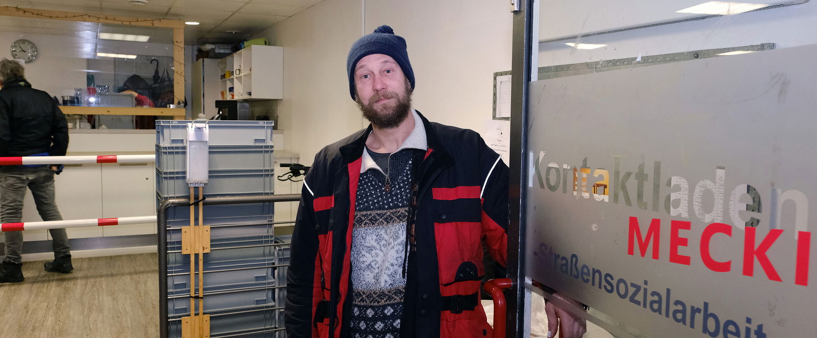 Eine als Mann lesbare Person mit Jacke und Mütze hält eine Tür mit Aufschrift Straßensozialarbeit auf.