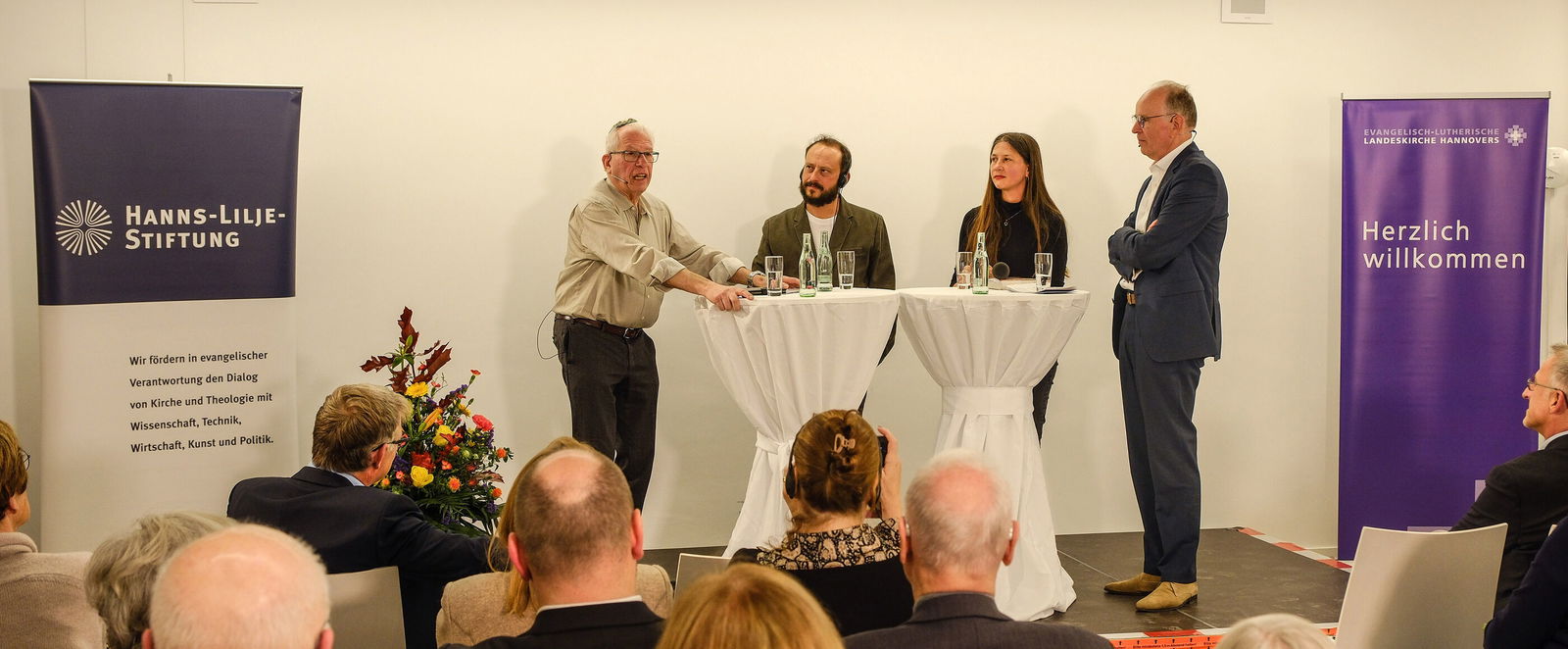 Drei männlich und eine weiblich zu lesende Person stehen an Stehtischen auf einem Podium und diskutieren.