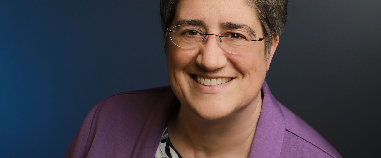 Eine weiblich gelesene Person mit Brille und grauen Haaren in Bluse und lila Jackett.