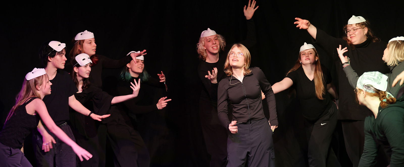 Eine Gruppe junger Menschen in schwarzer Kleidung auf einer Bühne, einige davon mit Maske.