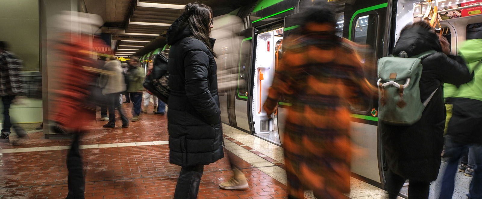 Eine Person steht still in einem U-Bahnhof, während alles um sie herum verschwommen wie im Zeitraffer zu erkennen ist: eine U-Bahn, andere Menschen.