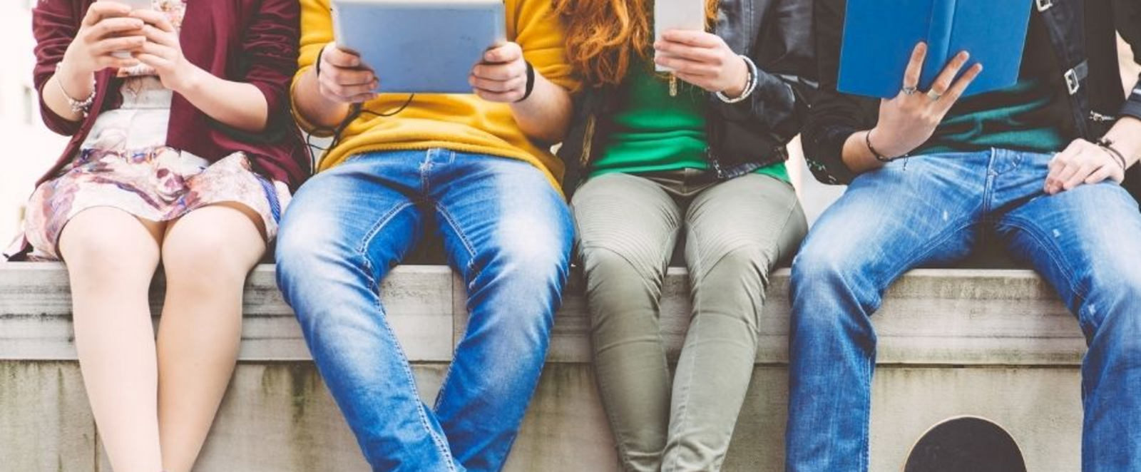 Vier junge Menschen sitzen auf einer Balustrade und haben Tablets, Handys und ein Buch in Händen.