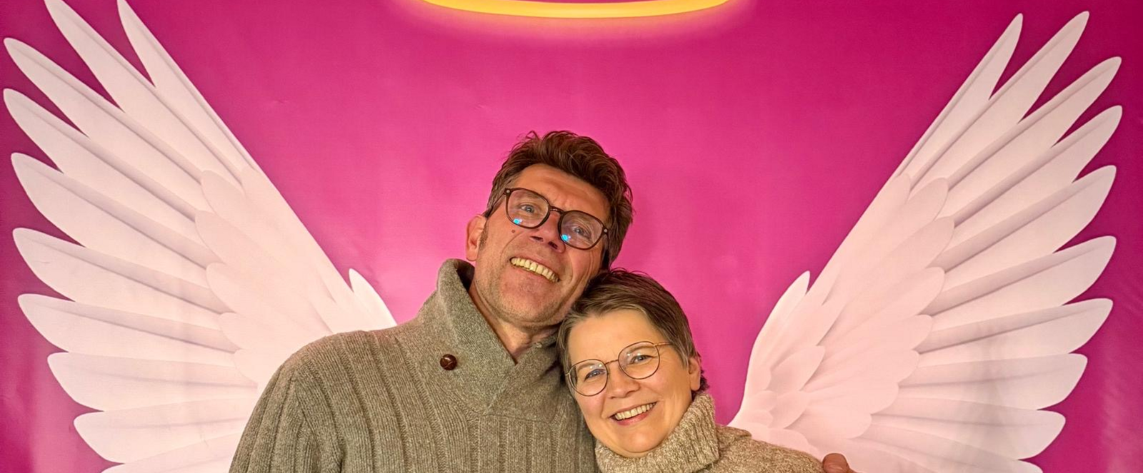 Eine männlich und eine weiblich gelesene Person halten sich im Arm, hinter ihnen eine Wand, auf der Engelsflügel und ein Heiligenschein gemalt sind.