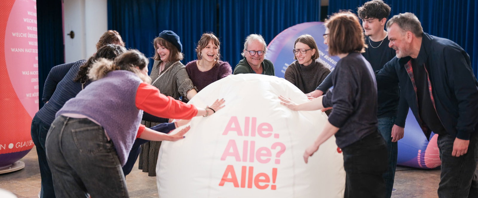 Mehrere Personen rollen einen großen Ball mit der Aufschrift: Alle. Alle? Alle!
