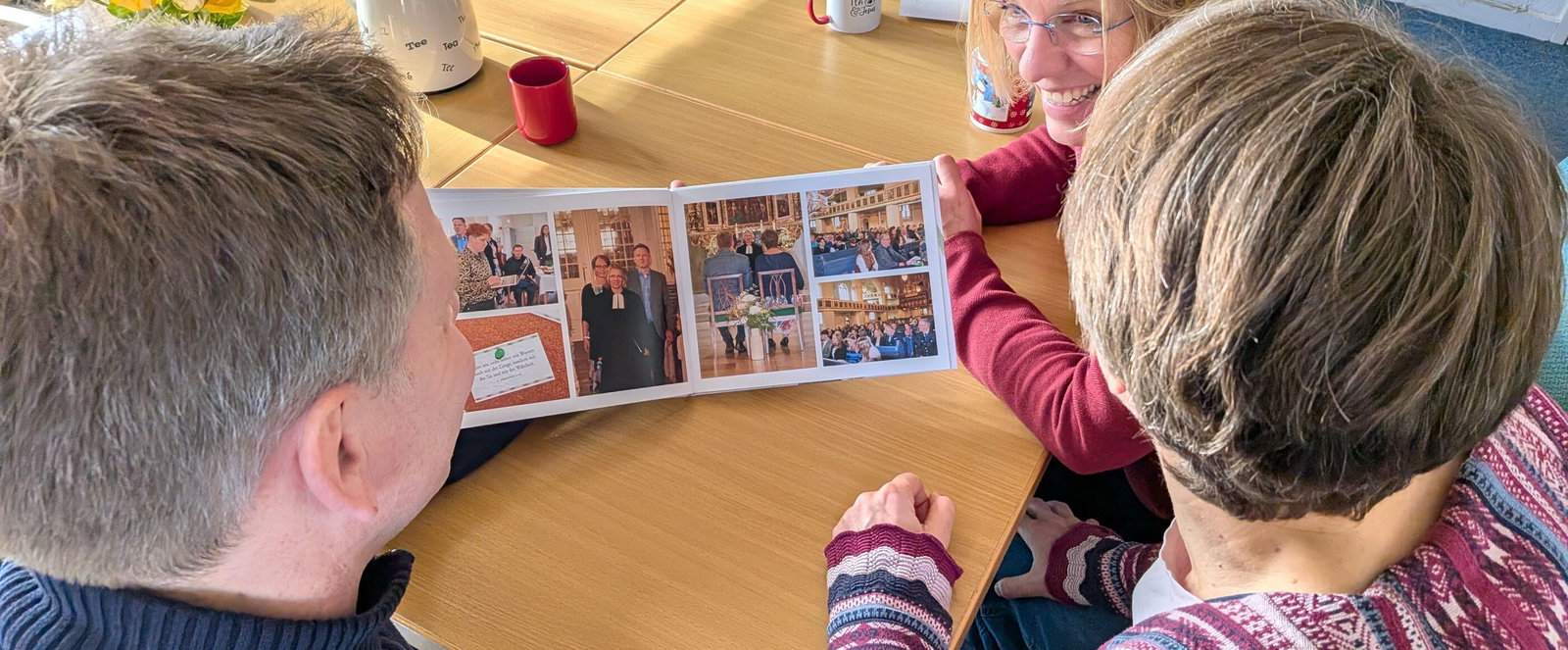 Drei Personen sitzen an einem Tisch und schauen in ein Fotobuch.