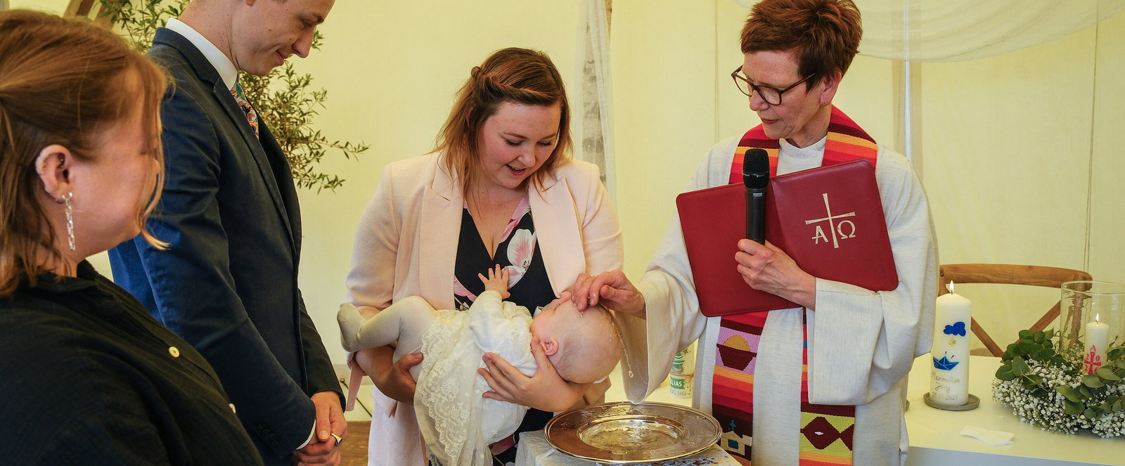 Eine Pastorin tauft in einem Zelt ein Baby, das die Mutter über die Taufschale hält.