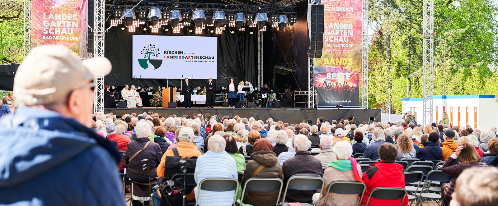 Eine große Menschenmenge sitzt vor einer Bühne, auf der ein Gottesdienst gefeiert wird.