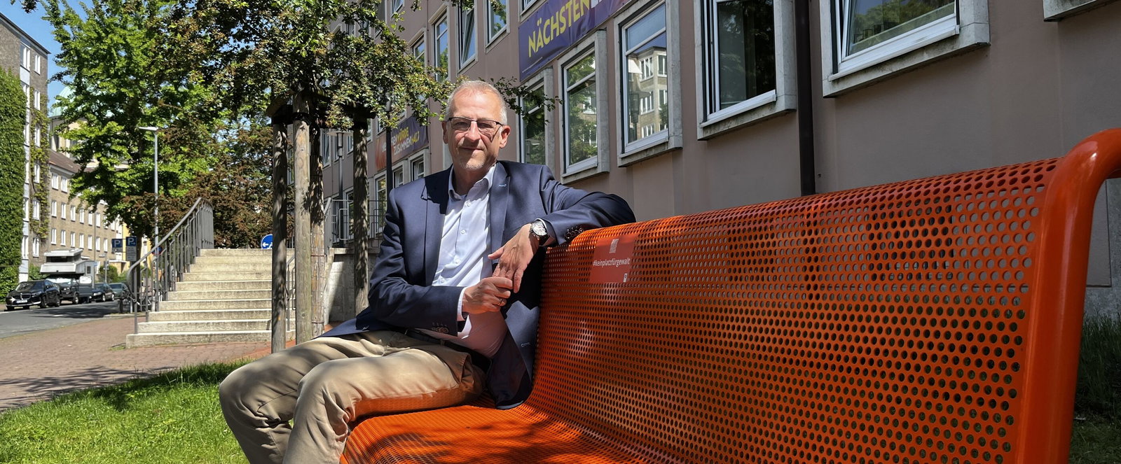 eine männlich gelesene Person sitzt auf einer orangefarbenen Bank draußen vor einem Gebäude