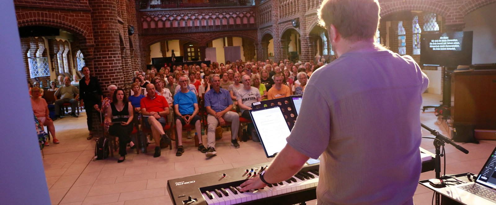 Eine als Mann lesbare Person steht am Keyboard und singt mit vielen Menschen in einer Kirche.