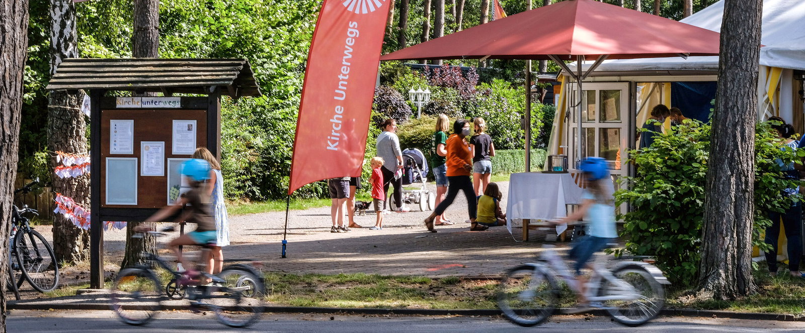 Eine Beachflag mit der Aufschrift „Kirche unterwegs“ auf einem Campingplatz. Im Vordergrund fahren Kinder auf einem Fahrrad.