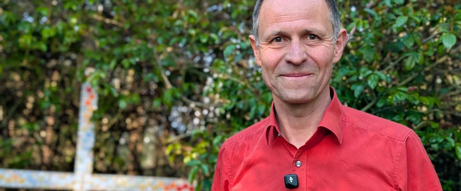 Eine als Mann lesbare Person mit rotem Hemd vor grünen Sträuchern, neben ihm ein angemaltes Holzkreuz.