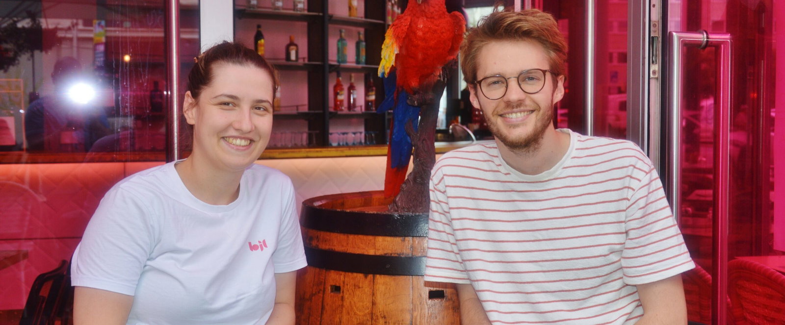 Links ist eine Frau und rechts ein Mann vor einer Bar im Hintergrund.