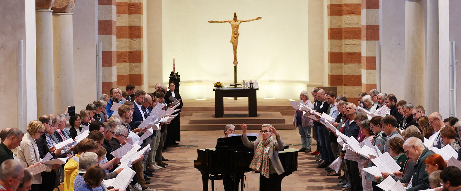 Viele Menschen singen in einer Kirche. Eine als Frau lesbare Person dirigiert, eine andere spielt am Flügel.