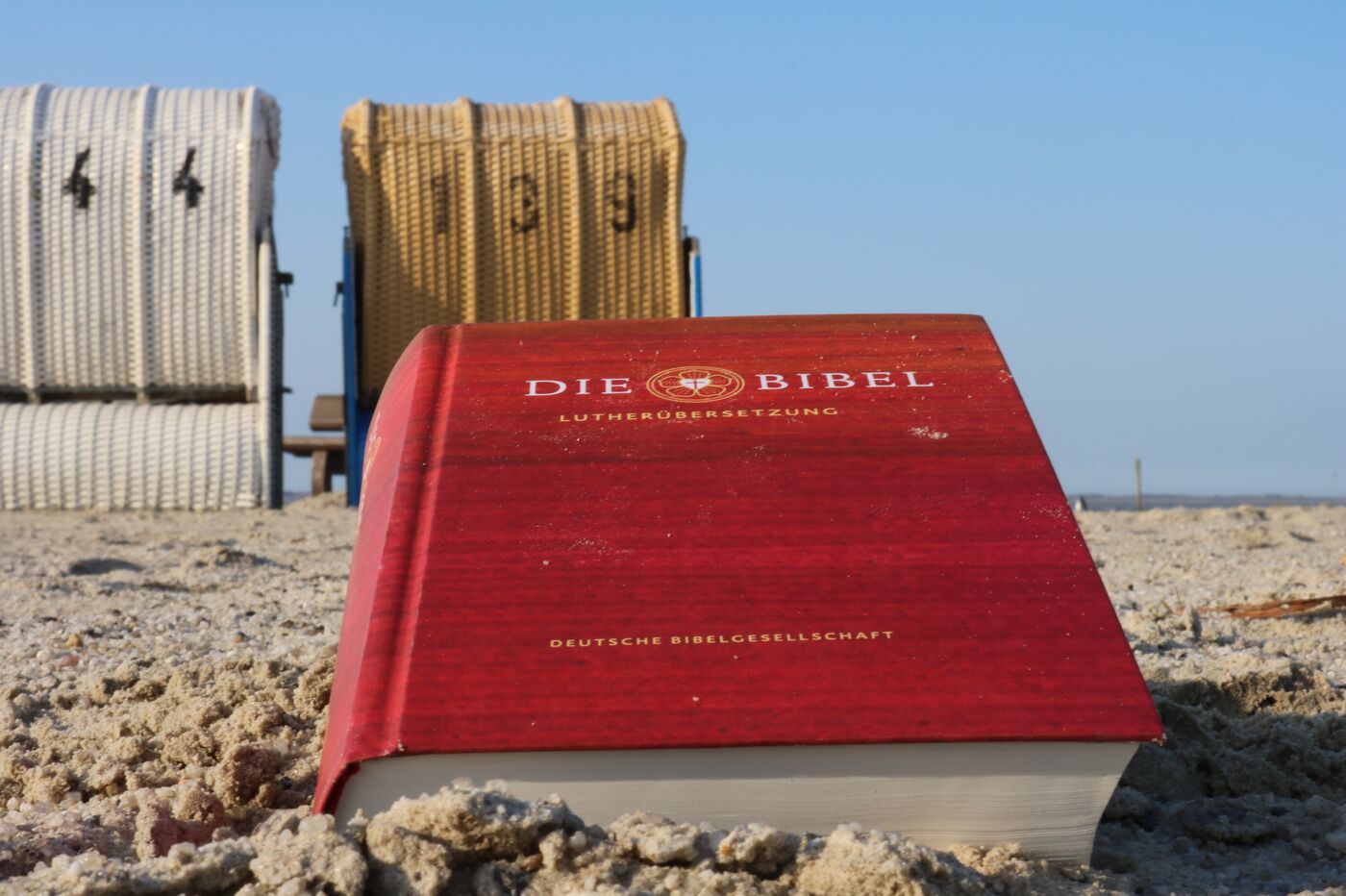 Eine Bibel im Sand am Strand. Im Hintergrund sind Strandkörbe zu erkennen. In der Urlaubsseelsorge kommt die Gute Nachricht Gottes natürlich auch an den Strand.