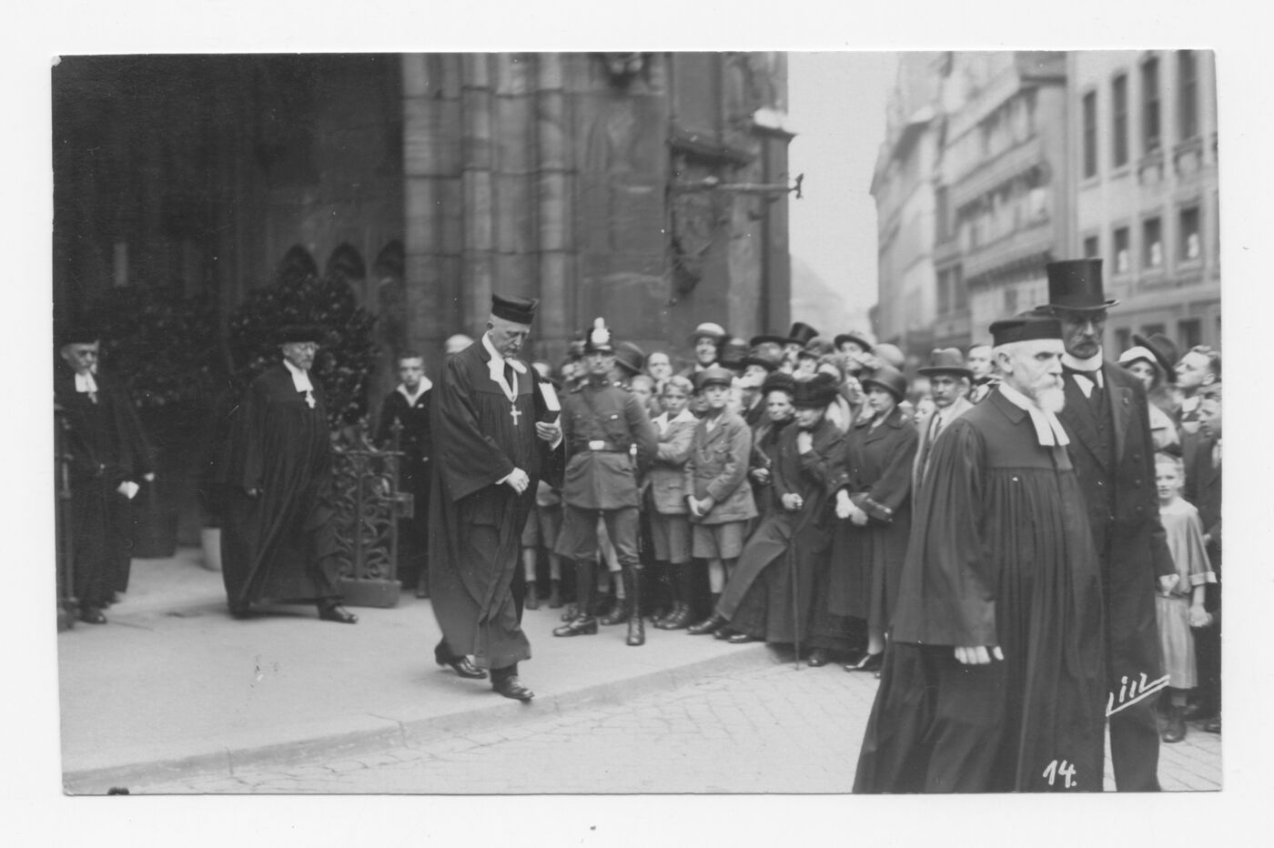 Das Bild zeigt eine Schwarzweiß-Aufnahme mit einer männlich lesbaren Person, die im Talar die Treppenstufen vor einer Kirche heruntergeht. Neben der Treppe wartet auf dem Vorplatz eine große Gruppe von winterlich angezogenen Menschen.