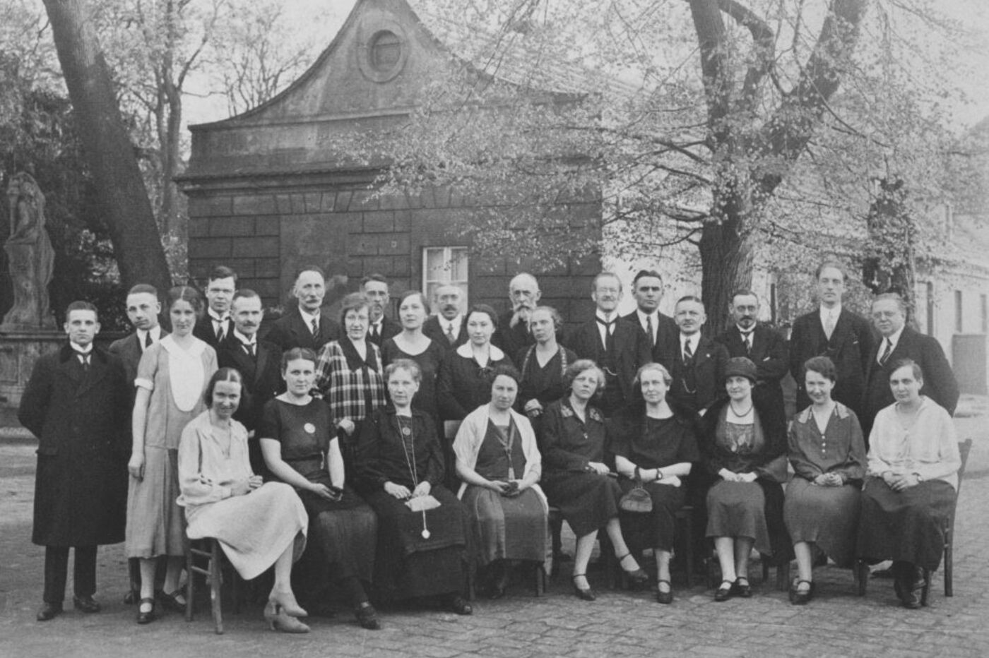 Das Bild zeigt eine alte Schwarzweiß-Aufnahme mit einer Gruppe von männlich und weiblich lesbaren Personen, die sich zu einem Gruppenbild im Park aufgestellt haben.