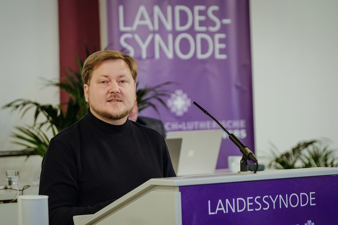 Eine männlich gelesene Person mit kurzen blonden Haaren und Vollbart hinter einem Redepult